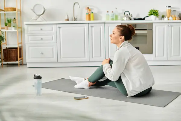 stock image Middle aged woman peacefully practices yoga on her mat in a cozy kitchen.