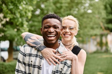 Neşeli bir Afrikalı Amerikalı adam beyaz bir kadını kollarında tutuyor, dışarıda parkta bir aşk anını paylaşıyor..