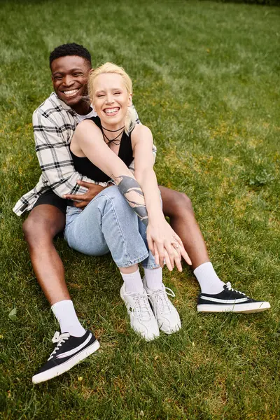 stock image A multicultural couple, an African American man and a Caucasian woman, sitting contently on the grass in a park.