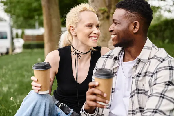 A multicultural couple, an African American man and Caucasian woman, enjoying coffees while seated on lush green grass in a park.