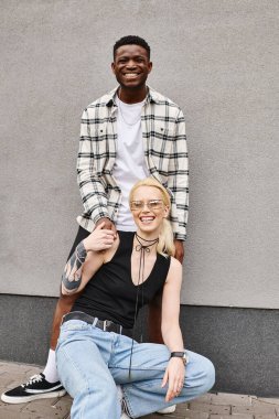 Farklı etnik kökenlere sahip bir erkek ve kadın, şehir ortamında gri bir binanın yanında birlikte oturuyorlar..