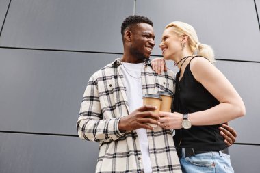 A joyful multicultural couple, consisting of a man and a woman, stand close together in front of a grey urban building. clipart
