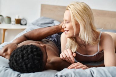 A sensual moment captured as a multicultural man and woman embrace intimately while laying on a bed at home. clipart