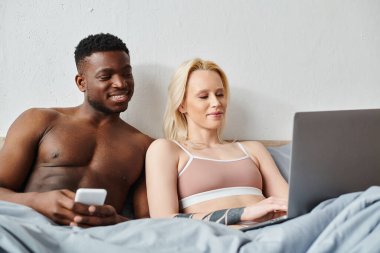 Multicultural couple sitting on bed, engrossed in laptop screen. clipart