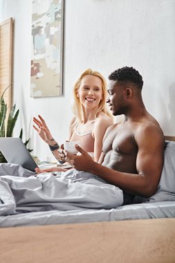 Seksi, çok kültürlü bir çift, erkek arkadaş ve kız arkadaş, yatakta oturuyor, birlikte dizüstü bilgisayara odaklanmış..