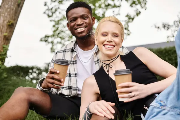 A happy multicultural couple, an African American man and a Caucasian woman, sitting on grass holding coffee cups.