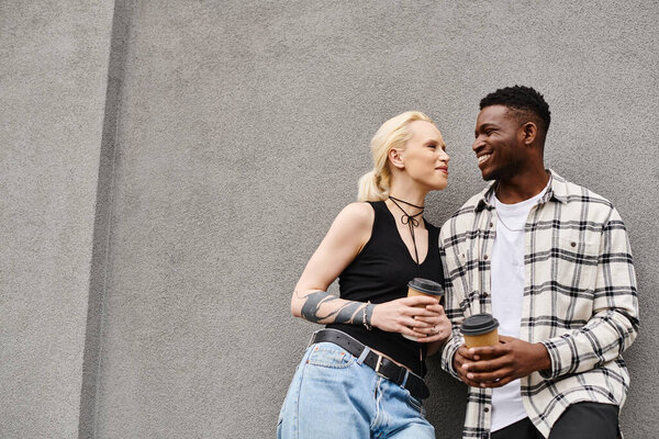 A happy multicultural couple, a man and a woman, standing close together on an urban street near a grey building.