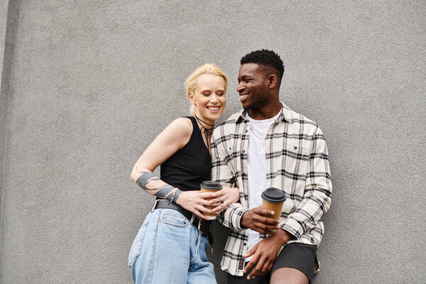 A happy, multicultural couple, a man, and a woman, stand closely together on an urban street near a grey building.