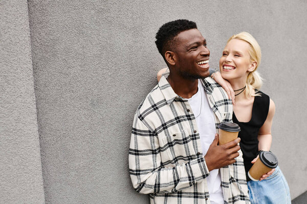 A multicultural couple, a man and a woman, stand happily together on an urban street next to a grey building.