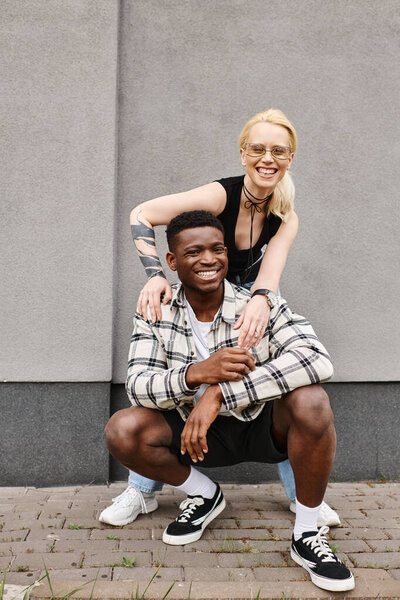 A joyful multicultural boyfriend and girlfriend pose together on an urban street near a grey building, smiling for the camera.