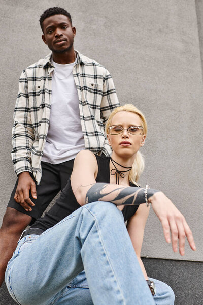 A man and a woman, a happy multicultural couple, sit together on the ground near a grey urban building, sharing a peaceful moment.