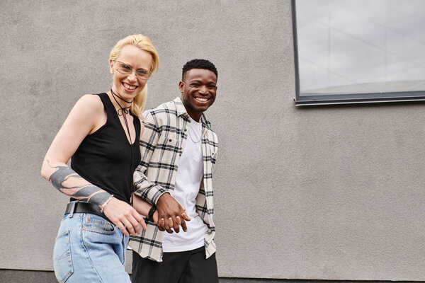 A happy multicultural couple, a man and a woman, standing together on an urban street near a grey building.