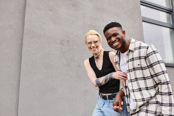Multicultural boyfriend and girlfriend stand happily together on an urban street near a grey building.