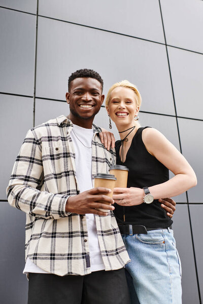 A multicultural couple, a man and a woman, stand happily side by side on an urban street near a grey building.