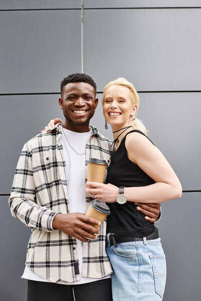 A happy, multicultural couple stands arm in arm on an urban street near a grey building, radiating joy and togetherness.