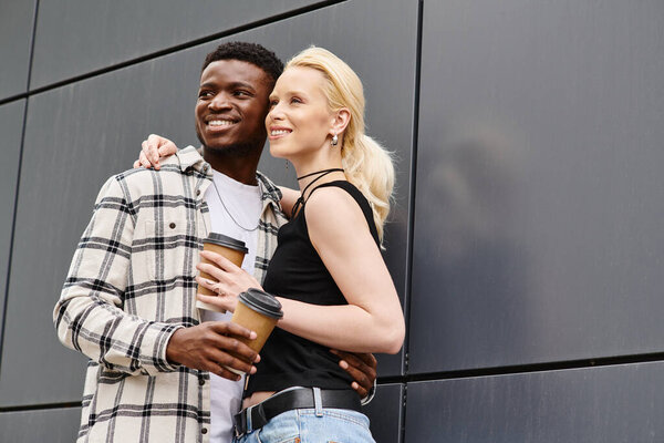 A happy, multicultural couple standing next to each other on an urban street near a grey building.
