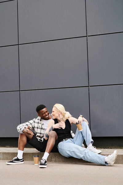A happy multicultural couple sitting beside each other on the ground near a grey urban building, sharing a quiet moment.