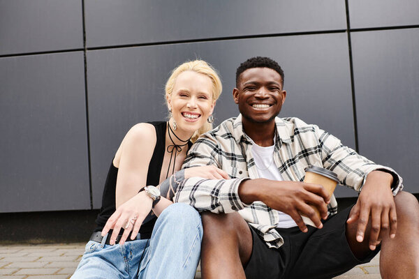 A happy multicultural couple sitting on the ground near a grey building in an urban street, sharing a moment of togetherness.