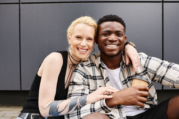 A happy, multicultural couple - boyfriend and girlfriend - sitting next to each other on an urban street near a grey building.