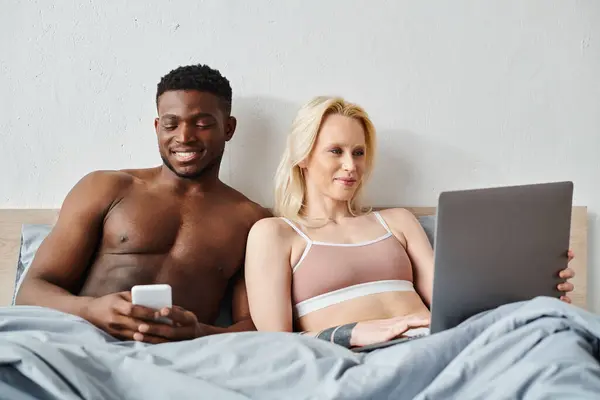 A multicultural couple sits on a bed, engrossed in a laptop screen together.