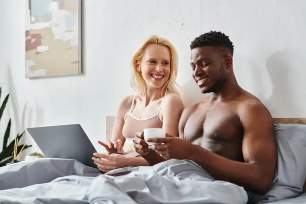 stock image A multicultural boyfriend and girlfriend sit on a bed, captivated by the screen of a laptop.