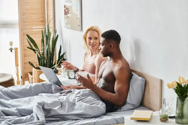 A multicultural boyfriend and girlfriend sit on a bed, engrossed in a laptop screen.
