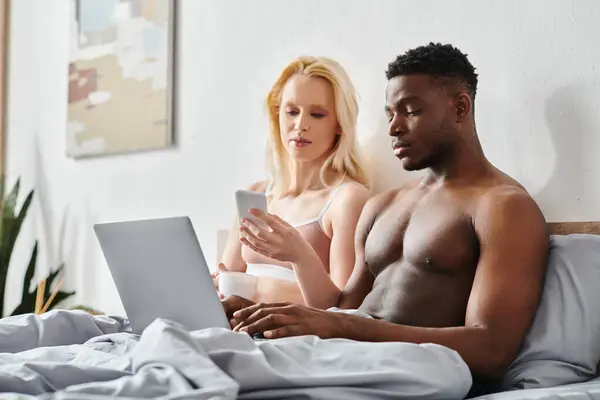 A multicultural boyfriend and girlfriend sit on a bed, intensely focused on a laptop screen.