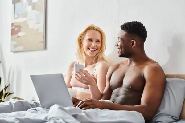 A multicultural couple, a man and woman, sit on a bed together, engrossed in the screen of a laptop.