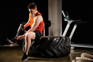 Protez bacağı olan engelli bir adam spor salonunda egzersiz yaparken lastiğe güvenerek oturuyor..