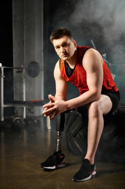 Bir spor salonunda siyah bir lastiğin tepesinde oturan protez bacaklı bir adam güç ve kararlılık gösteriyor.