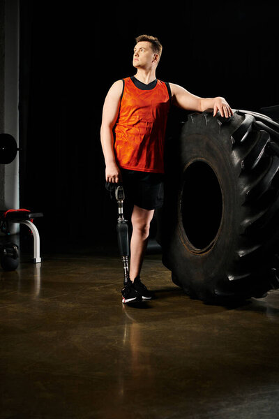 A man with a prosthetic leg standing next to a massive tire in a gym.