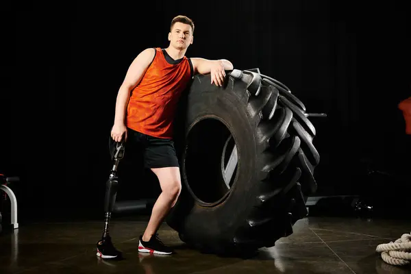 在体育馆里 一个有假腿的男人自信地站在一个巨大的轮胎旁边 展示了他的力量和决心 — 图库照片