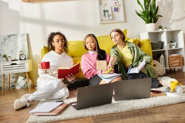 Üç ırklar arası genç kız rahat bir kanepede kitap ve dizüstü bilgisayarlarla ders çalışıyor, arkadaşlık ve eğitimi somutlaştırıyor..