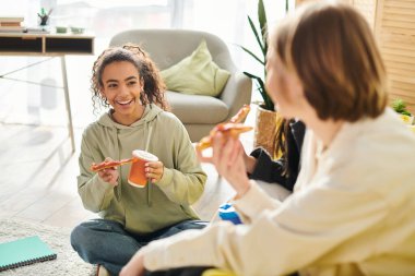 Two interracial teenage girls sitting on the floor, eating pizza and enjoying each others company in a cozy setting. clipart