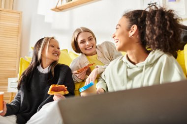 A diverse group of teenage girls laughing and chatting while sitting on a couch and eating delicious pizza together. clipart