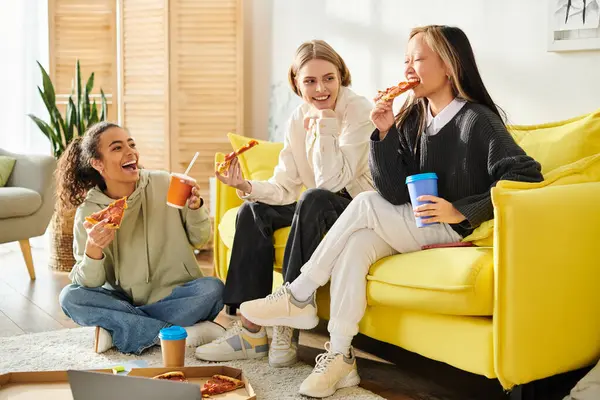 stock image Three diverse young women enjoy pizza and coffee on a cozy couch, bonding over food and laughter.
