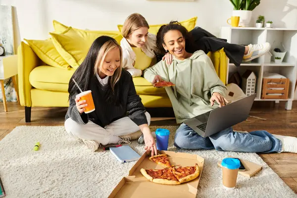Farklı etnik kökenlere sahip üç genç kız yerde rahatça oturuyor, pizza dilimlerinin tadını çıkarıyor ve içkilerini yudumluyor..