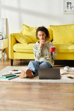 A young woman sits on the floor, gracefully clutching a warm cup of coffee while studying on her laptop at home. clipart