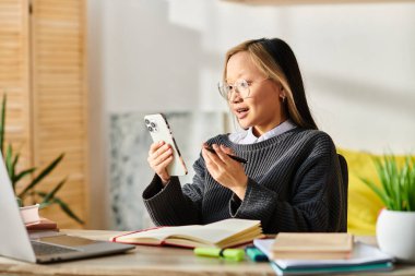 Genç bir Asyalı kadın e-öğrenmeye dalmış, bir masada oturuyor ve öğrenim kaynakları için cep telefonuna bakıyor..