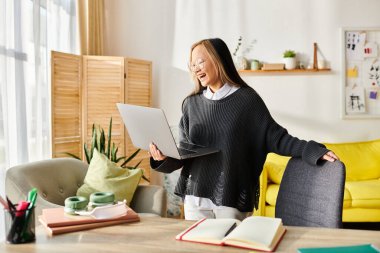 Genç Asyalı bir kız, elinde bir dizüstü bilgisayarla oturma odasında kendini derslerine kaptırmış bir şekilde duruyor..