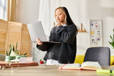 Genç bir Asyalı kadın dizüstü bilgisayarın önünde duruyor, evde e-öğrenimle uğraşıyor..