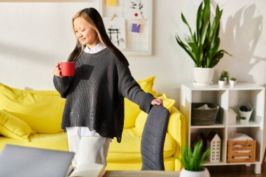 A young Asian woman standing in a living room, enjoying a cup of coffee while e-learning with her laptop. clipart