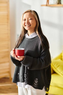 Genç bir Asyalı kız evde e-öğrenirken neşe içinde bir fincan kahve tutuyor..