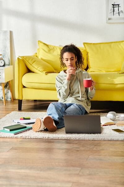 A young woman sits on the floor, gracefully clutching a warm cup of coffee while studying on her laptop at home.