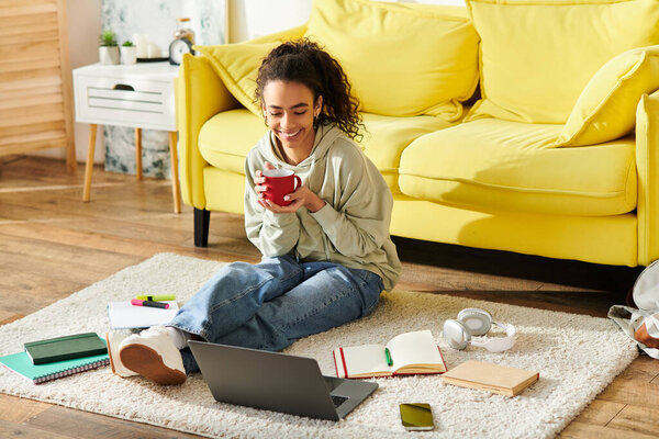 A teenage girl sits on the floor, laptop nearby, enjoying a cup of coffee.