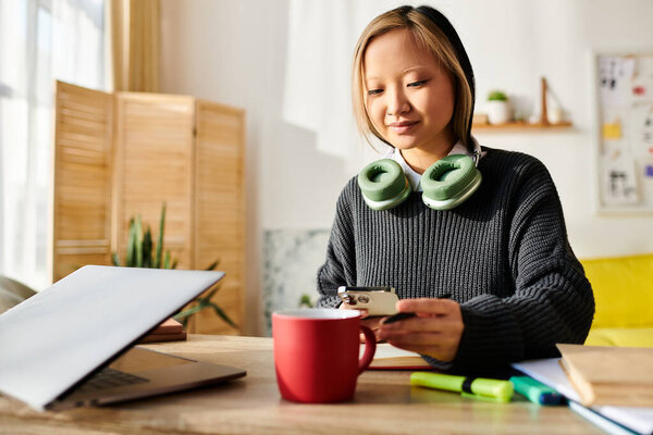 A young Asian woman sits at a table, savoring a cup of coffee while studying with a laptop at home.