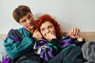 Canlı moda kıyafetleri içinde neşeli ve çekici bir çift evde yataktayken sarılıp gülüyor.