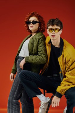 Parlak giyinmiş, güneş gözlüklü, şık ve çekici bir adam. Sandalyede güzel bir kız arkadaşın yanında oturuyor.