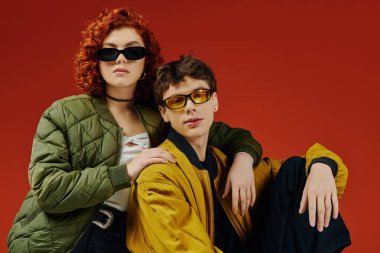 Güneş gözlüklü, kırmızı arka planda birlikte poz veren şık giyinen çekici bir çift.