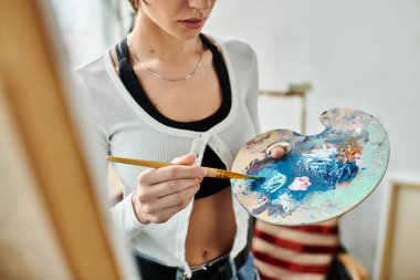 A woman delicately holds a paintbrush and palette, immersed in creativity. clipart